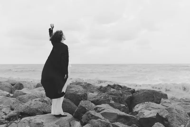 Woman stands on jagged rocks by the sea, with an outstretched arm, wearing a coat in a windy black and white landscape, evoking solitude and resilience.