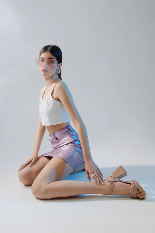 Young woman in a white crop top and shiny pink skirt poses kneeling on the floor. She wears sunglasses and high heels. Concept for woman beauty fashion and style.