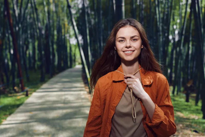 Woman smile portrait walking on bamboo forest path outdoors in orange jacket, young adult enjoying nature, serene stroll with sunlight through trees, casual happy relaxed expression