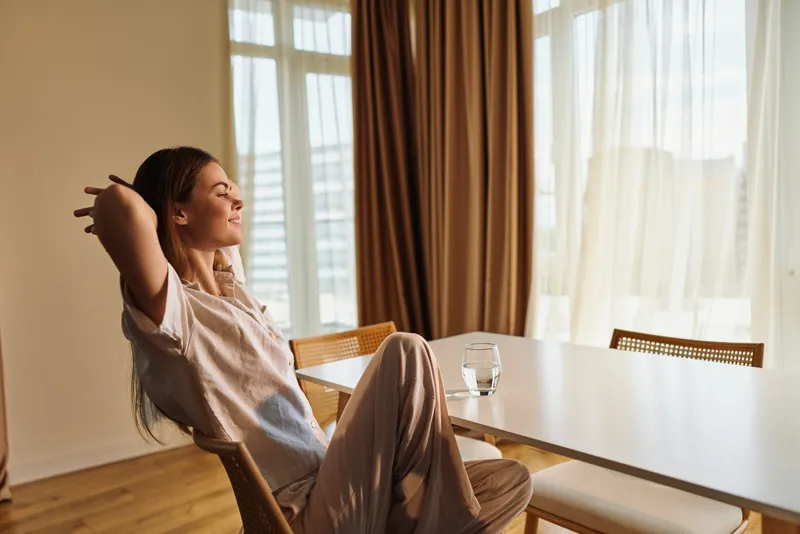 Woman relaxation home sunlight chair table stretching morning portrait of a young female smiling in sunlit dining room, casual outfit, peaceful comfort and cozy domestic lifestyle.