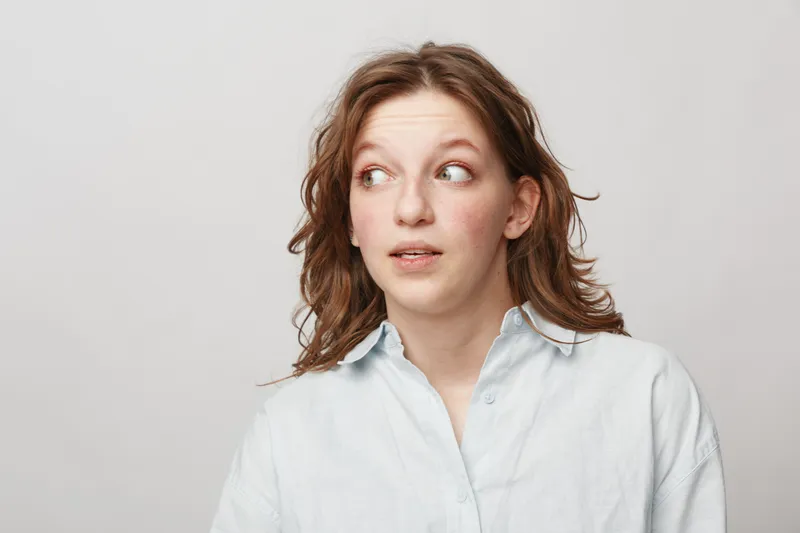Young woman with curly hair wearing white shirt looks sideways with curious expression on light background. Concept of surprise, doubt, and casual lifestyle portrait.