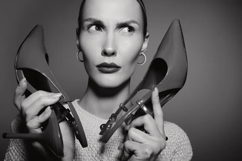 White woman with light skin holds two high heel shoes near face in black and white studio portrait.