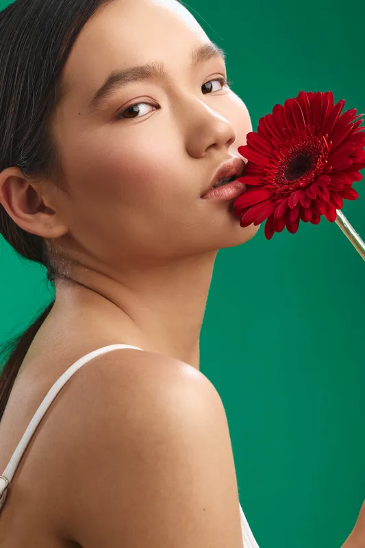 Asian woman poses with a red flower against a vivid green backdrop, creating a graceful portrait that highlights beauty, calm, and refined style.