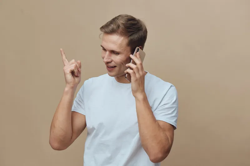 Young smiling man talking on mobile phone wearing casual white t-shirt indoors isolated on beige background concept communication technology lifestyle