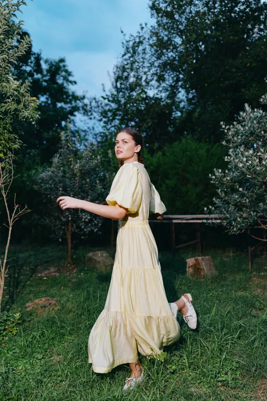Woman in a flowing yellow dress walks through a garden, capturing a carefree outdoor fashion moment with soft grass and trees in the background.
