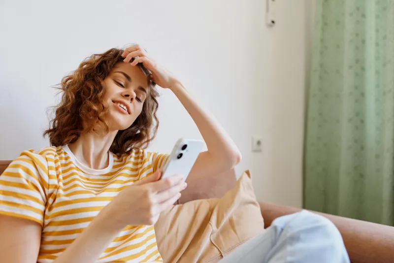 Casual woman at home sits on a sofa, smiling while using a smartphone, relaxed mood, bright natural light, cozy living room, lifestyle portrait, simple decor