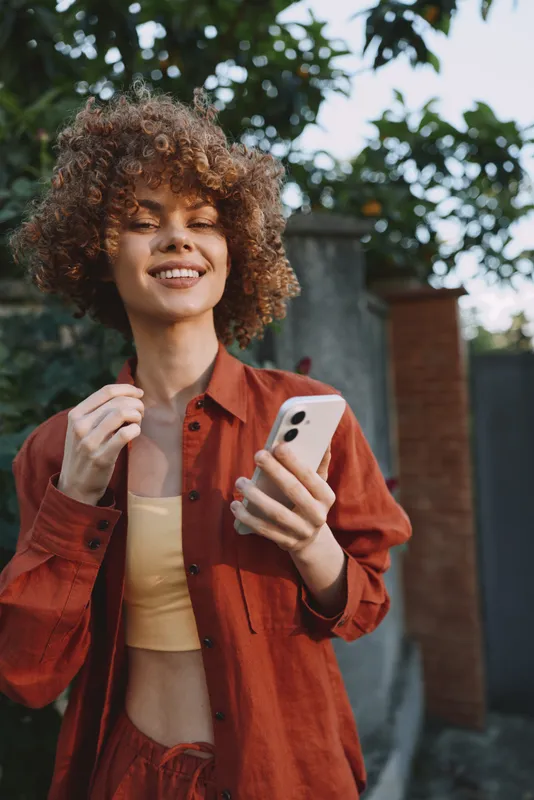 A smiling woman in a casual red shirt stands outdoors, holding a smartphone. Bright natural light highlights her relaxed vibe. Concept for lifestyle, fashion, and portrait storytelling
