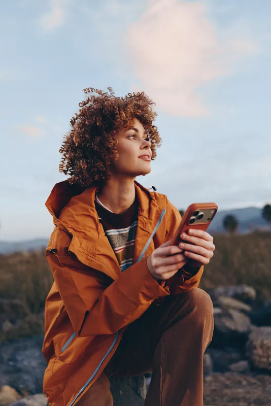 Woman lifestyle outdoors, curly haired woman in orange jacket, holding smartphone, gazing at screen, bright sunlight, open sky, natural landscape, calm relaxed pose, quiet breeze