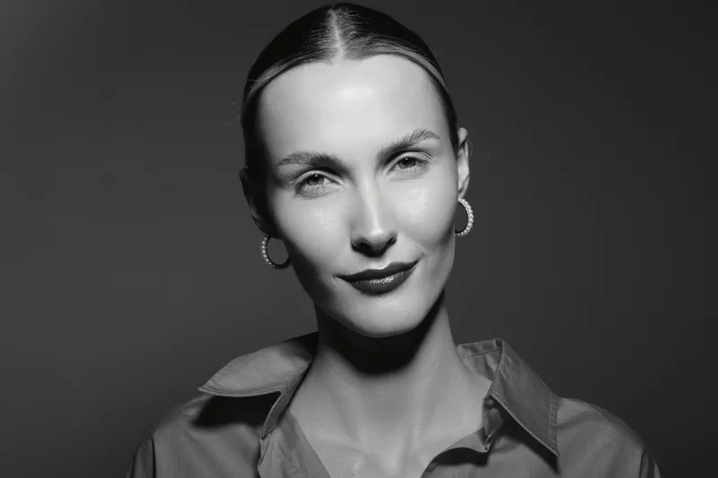 Portrait of a Caucasian woman with pale skin and slicked back hair wearing hoop earrings and a collared shirt. Studio lighting creates a confident and elegant mood.