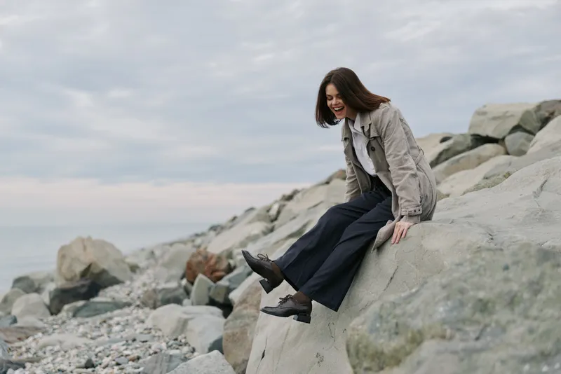 Young woman in casual outfit sits on large rocks near the sea, smiling and enjoying the fresh air. Cloudy sky and rocky shore create a peaceful outdoor atmosphere.