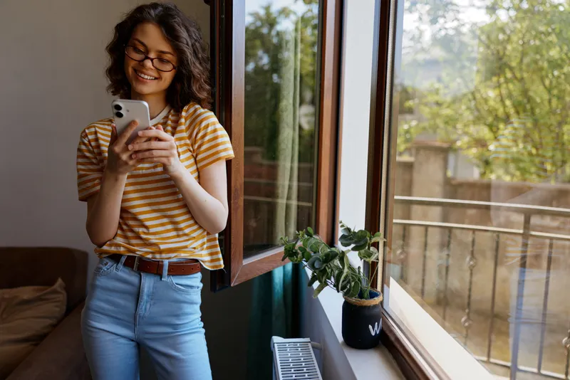 Young woman stands by a sunny window in a striped tee scrolling on her smartphone with a relaxed cheerful mood in a cozy home setting