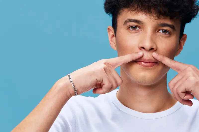 young man showing mustache gesture with fingers on blue background, casual white t-shirt, curly hair, playful expression, studio portrait with clean color contrast