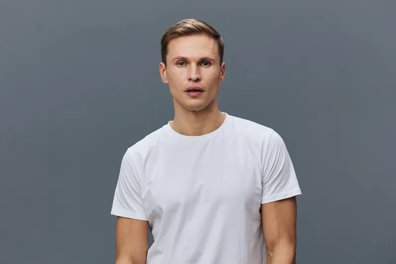 young caucasian man in white t-shirt looking surprised isolated on gray background close-up portrait mental health concept