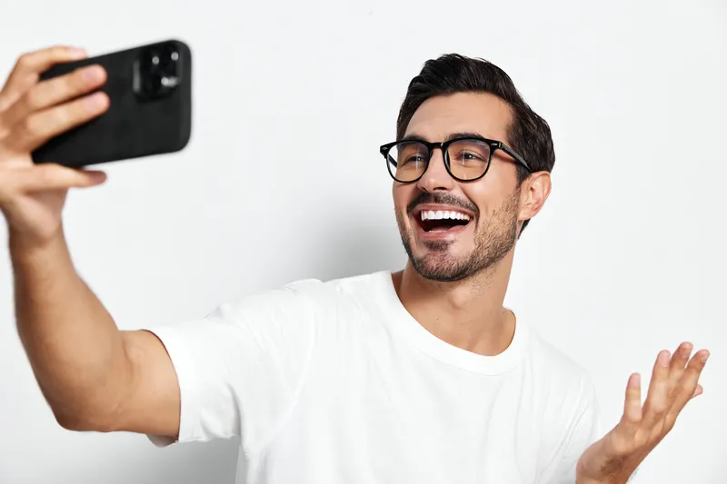 Happy smiling young man take selfie with smartphone, wearing stylish glasses and casual white shirt, looking cheerful and engaged, isolated on plain background.