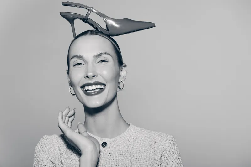 Smiling Caucasian woman with light skin wearing high heel shoe on head, posing in knitted sweater on white background. Concept of fashion and humor.
