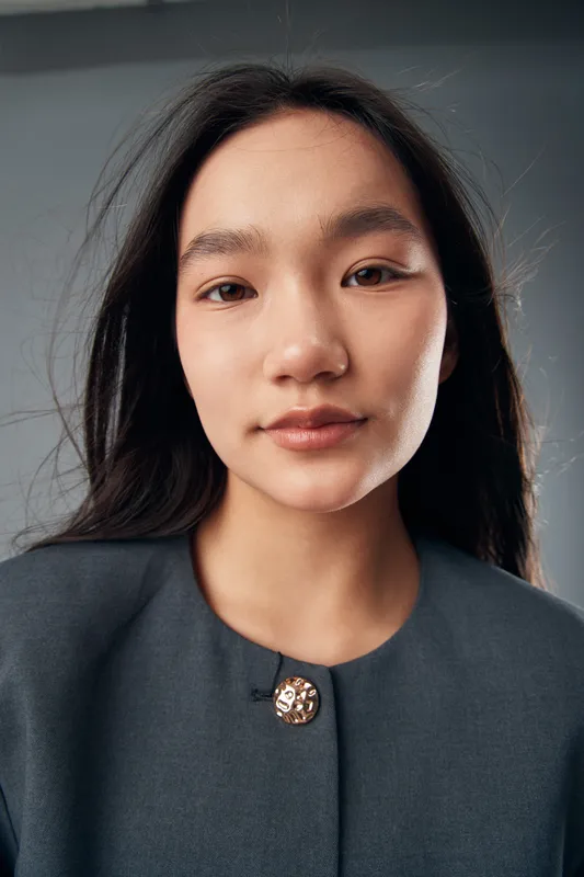Portrait of young woman with long black hair wearing a dark jacket looking at camera with neutral expression, minimal makeup and smooth skin in studio lighting setup.
