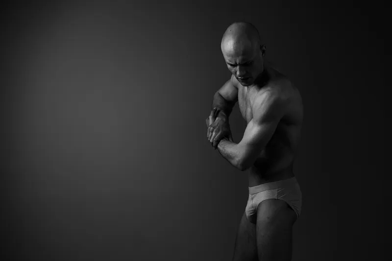 Black and white image of a muscular man posing thoughtfully, conveying strength and introspection with shadows highlighting his physique.