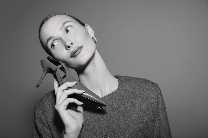 Young white woman with pale skin holding high heel shoe near face, looking up with thoughtful expression. Studio portrait in black and white against plain background.