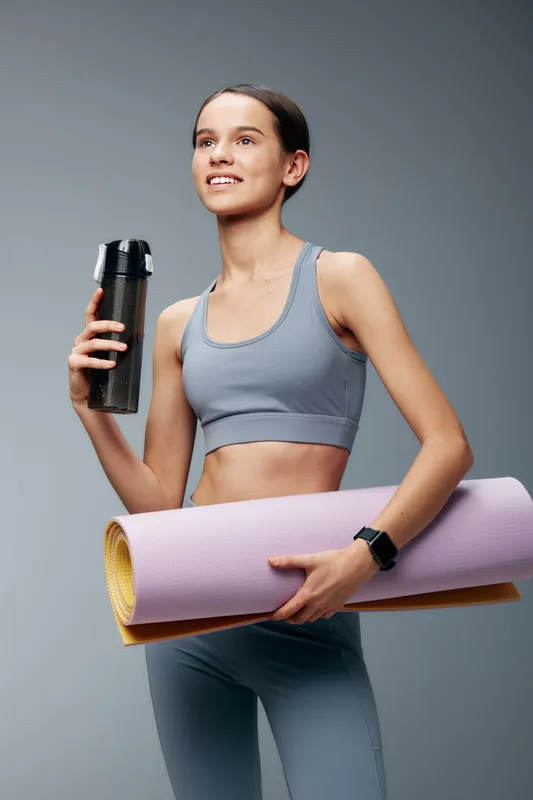 Female fitness enthusiast in athletic wear stands with a water bottle and rolled yoga mat, ready for a workout session in a clean studio.