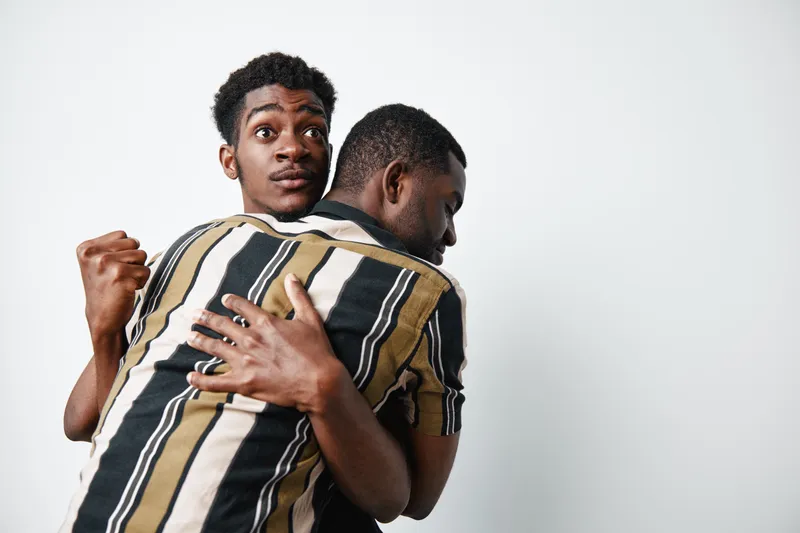 Two young black men with dark skin hugging each other in emotional support and friendship on white background. African ethnicity, casual clothing and close connection.