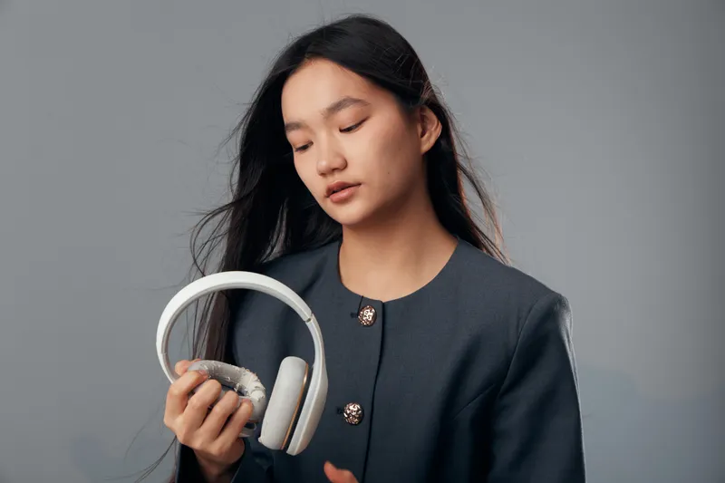 Young woman with long hair holding white wireless headphones looking down at earbuds in hand against gray background. Casual lifestyle, technology and music concept with focus on sound devices.