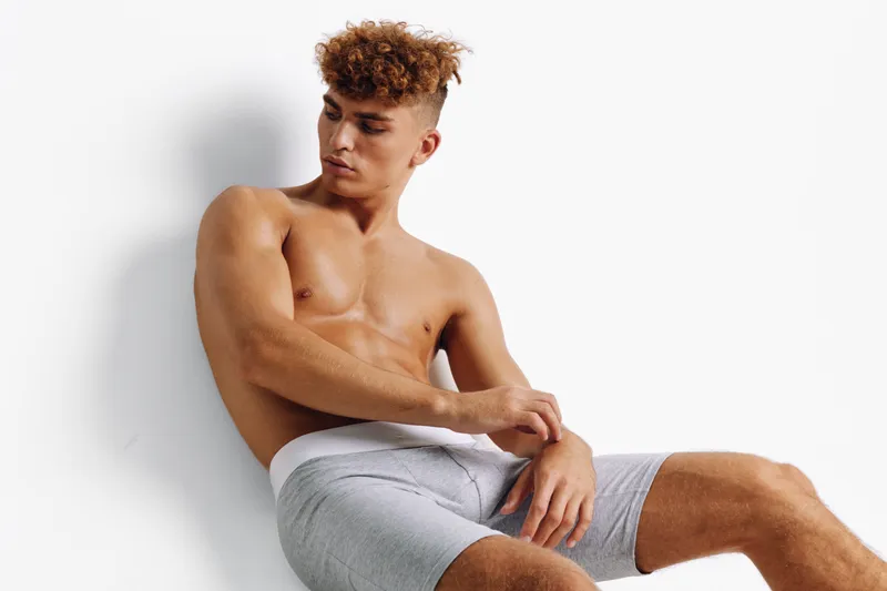 Young man with curly hair posing shirtless in gray shorts against white background, showing athletic build and relaxed expression in studio lighting.