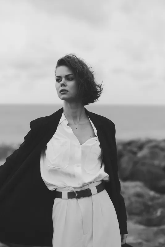 Fashion, model, outdoor, coastal, blazer, white shirt, elegance, wind, sea, dramatic visuals highlight a confident figure on a windy shoreline with a black coat open, minimalist styling and grayscale