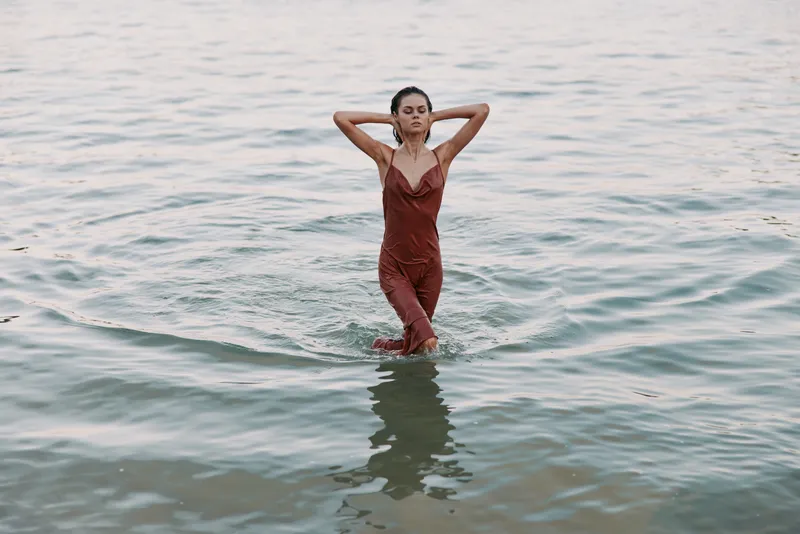 A confident woman stands waist deep in calm water, wearing a red swimsuit with hands behind her head, creating a striking pose against a serene seaside backdrop.