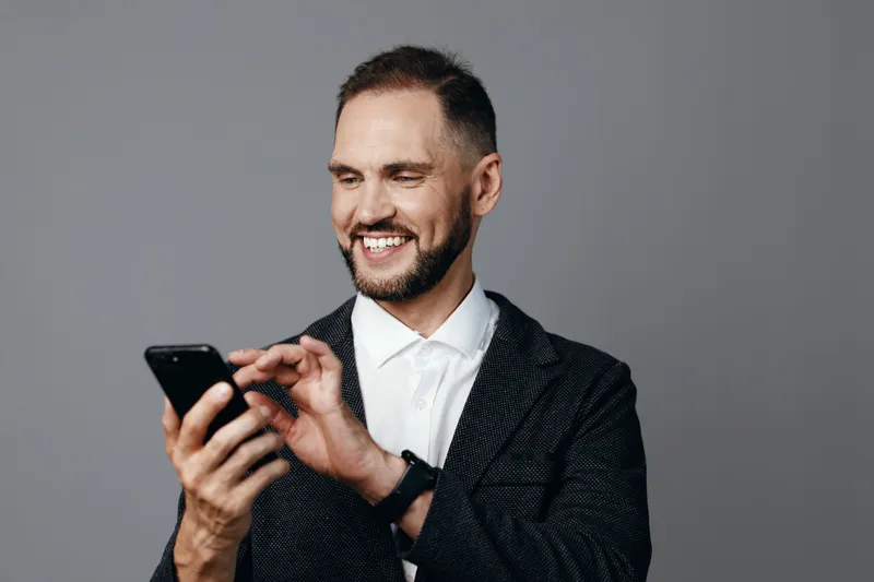 A professional businessman stands against a solid colored backdrop, smiling as he taps a smartphone with confident ease, conveying leadership, focus, and approachable expertise in a modern corporate