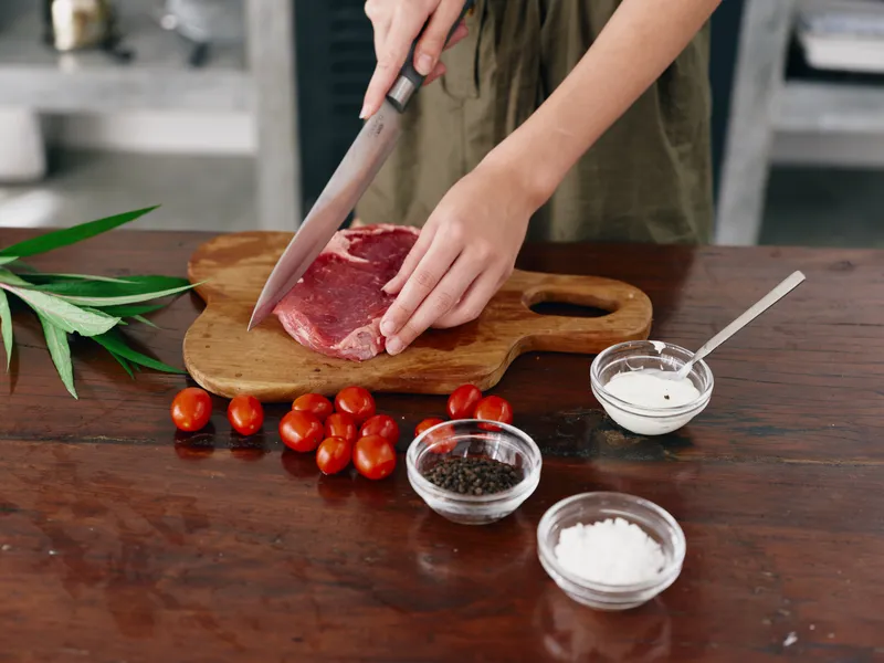 Женщина с ножом в руке нарезает мясо для стейка для жарки на кухне, на столе соль, перец и другие специи, красные помидоры черри и зелень, приготовление ужина