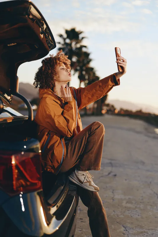 Woman lifestyle outdoor city street sunset scene, curly hair woman in jacket stands by open car door, holding phone up with hand, warm evening light, travel mood, candid portrait