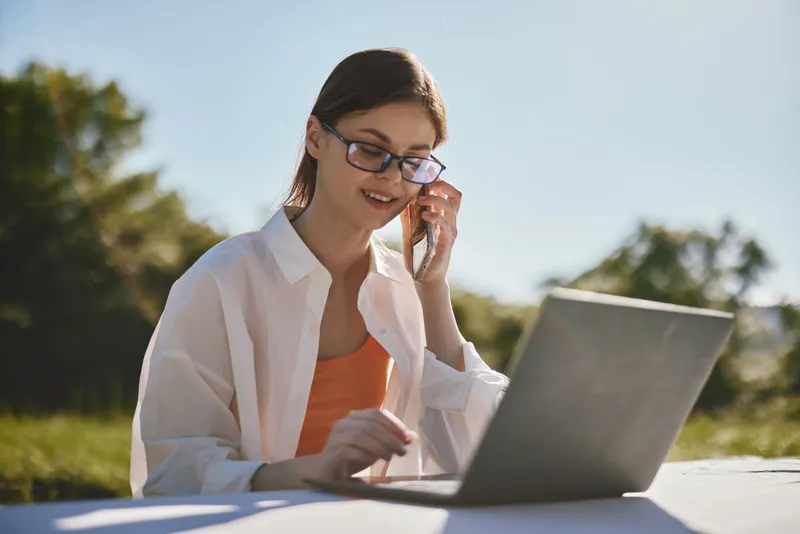 Young woman in glasses working on a laptop outdoors, wearing a casual white shirt and orange top, smiling while on the phone, showcasing a productive lifestyle