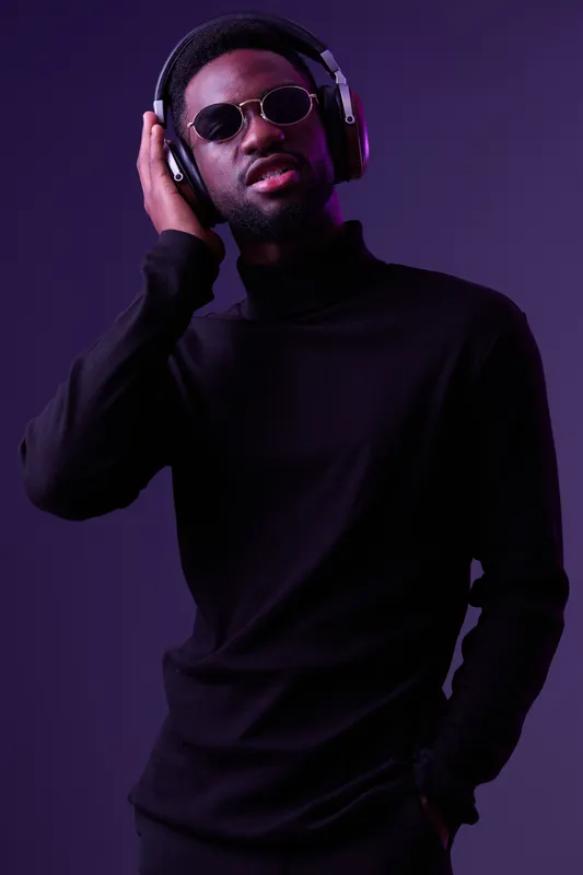 Stylish young man enjoying music with headphones, wearing a black turtleneck and sunglasses against a vibrant purple background