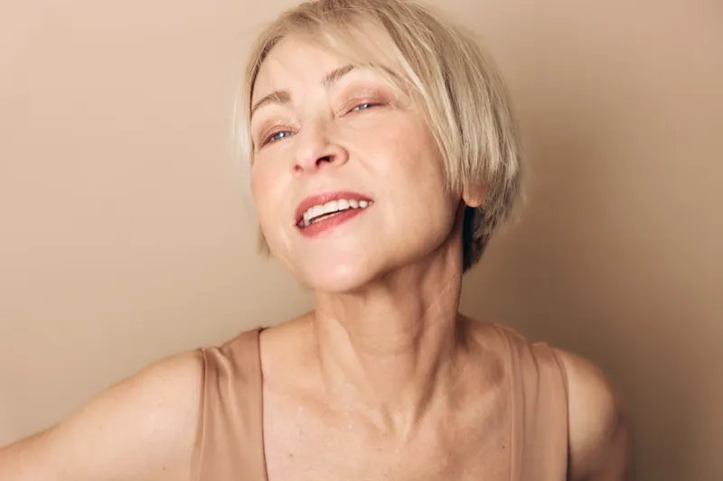 Smiling mature woman with short blonde hair wearing beige top poses against neutral background. Close-up portrait captures joyful expression and natural beauty with soft lighting.