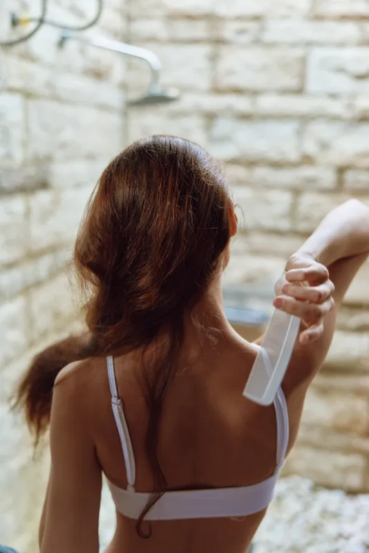 woman brushing hair in shower with stone wall background, long brown hair, white bra, intimate self-care moment, natural morning routine