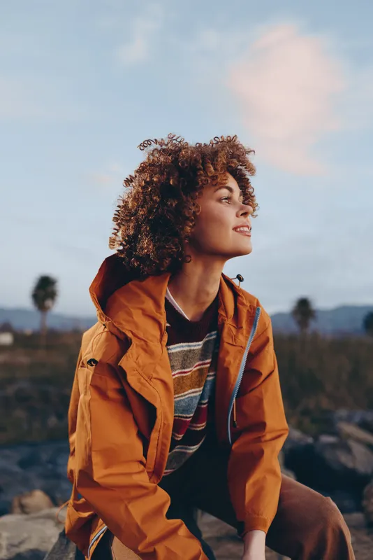 Woman curly hair outdoors portrait, orange jacket lifestyle, sunset sky nature background, warm soft light, wind in hair, relaxed smiling mood, everyday wellbeing, outdoor travel, fresh air