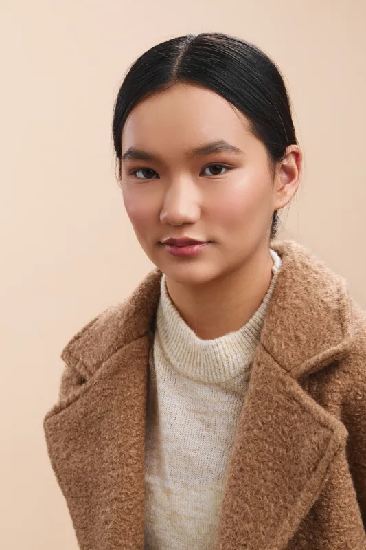 asian woman wearing a warm beige coat and soft sweater, posed in a light studio setting, presenting a calm, confident portrait with natural lighting and minimal background distractions.