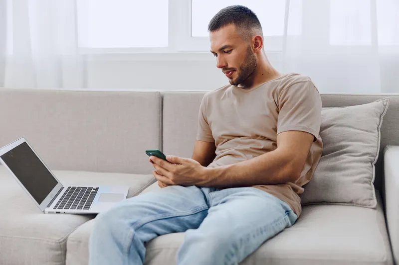 Focused man using smartphone while sitting on a cozy couch, surrounded by natural light and a laptop. Perfect for modern lifestyle and work-from-home themes.