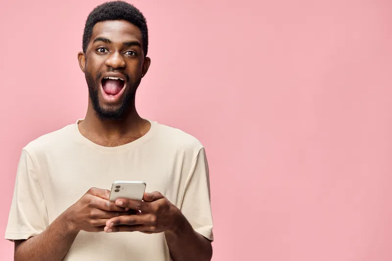 Joyful young African man holding a smartphone and smiling against a pink background, showcasing excitement and modern technology