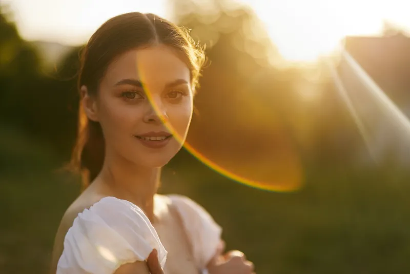 Beautiful young woman in a white dress smiling softly at sunset, surrounded by a warm, golden glow and natural greenery, capturing the essence of serene beauty