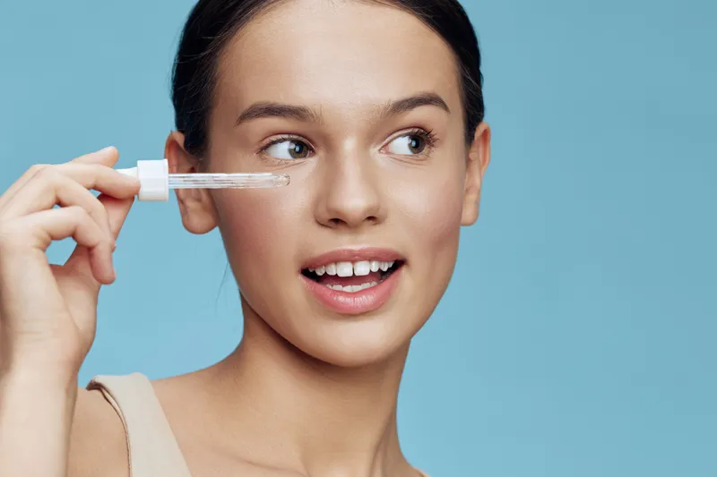 Young woman applying serum with dropper, showcasing clear skin and smiling, against a soft blue background, emphasizing beauty and skincare