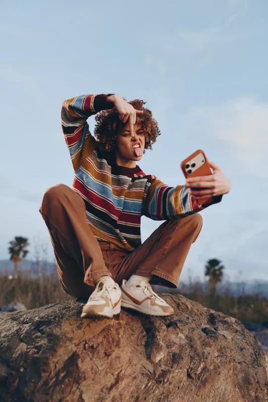 Woman takes a selfie outdoors, lifestyle moment on a rock, curly hair in sunlight, summer vacation mood, nature landscape behind, she wears casual sweater and jeans, relaxed smile