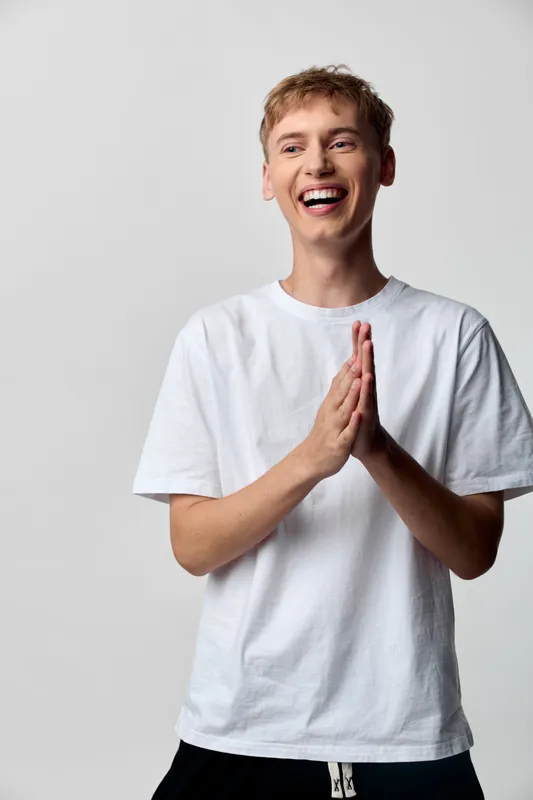 Joyful man with a bright smile and hands pressed in a prayer like gesture, casual white tee, studio style, warm and friendly mood, conveying gratitude and positive energy in daily moments