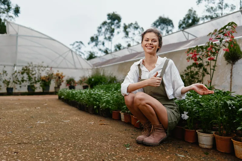 Happy young woman in a greenhouse showcasing vibrant plants, dressed casually in overalls and boots, emphasizing gardening and nature s beauty
