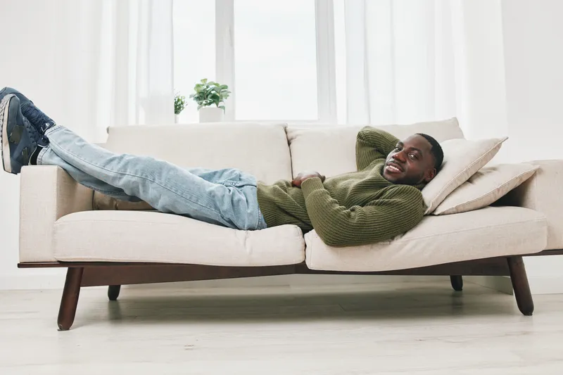 Young Black man relaxing on a comfortable sofa, smiling with joy while enjoying indoor leisure time in a cozy living room environment
