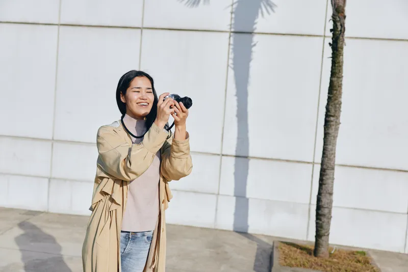 Woman with camera capturing a cheerful moment outdoors beside a white wall and palm tree, wearing a beige coat and denim, showcasing candid lifestyle photography in daylight