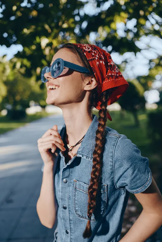 Lifestyle woman in modern boho-western denim street style wearing red bandana and sunglasses, smiling outdoors with film color effect under leafy trees.