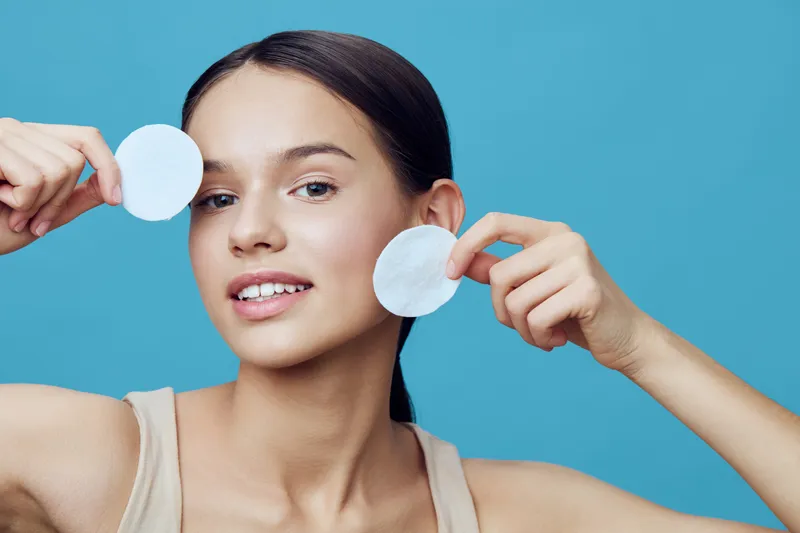 Young woman with a natural look holding cotton pads on a blue background, promoting skincare and self care routines for glowing skin