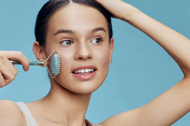 Young woman using a facial roller for skincare, showcasing radiant skin and a joyful expression against a soft blue background