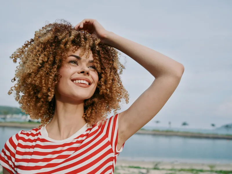 Young woman with voluminous curly hair wears a red and white striped shirt and smiles warmly at the camera on a sunny beach, exuding joy and carefree confidence.